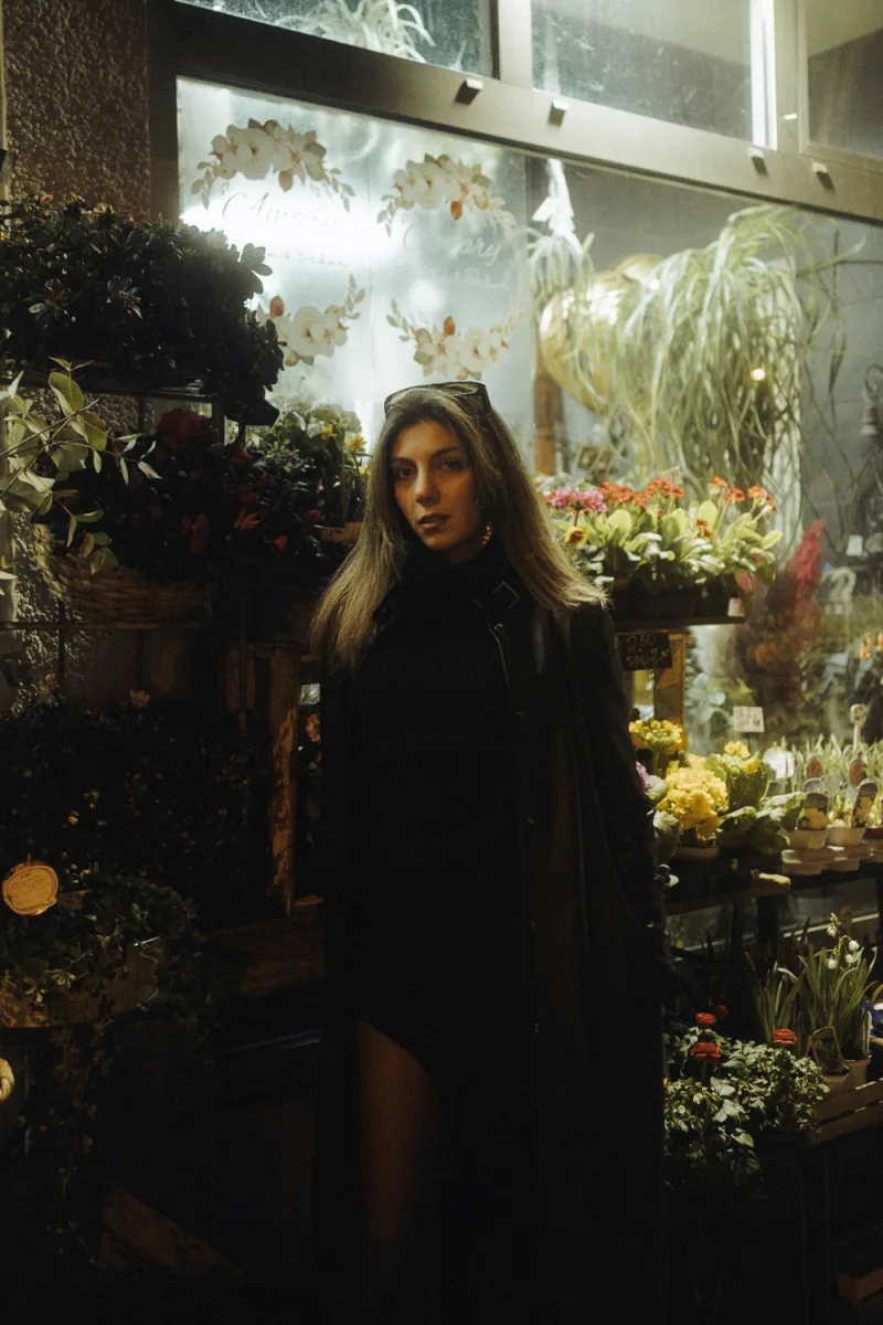 Woman with long dark hair surrounded by flowering plants inside illuminated shop