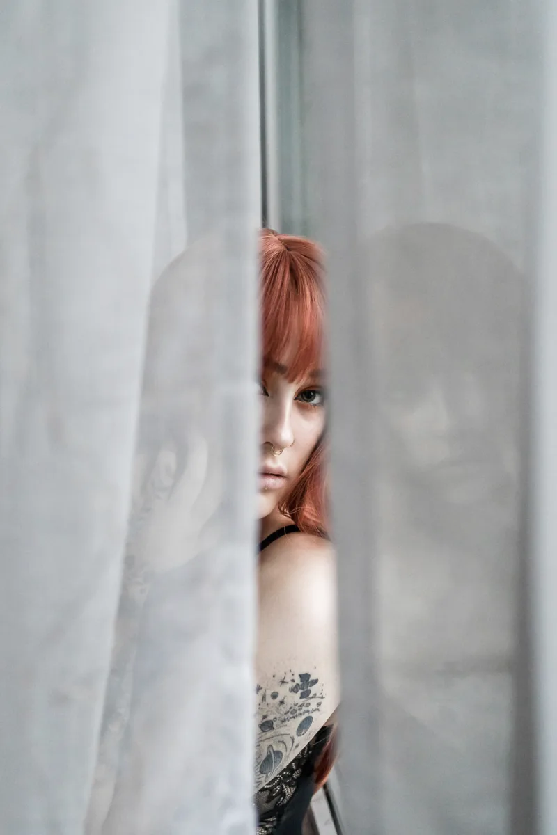 Woman with orange hair peering between white panels showing arm tattoo
