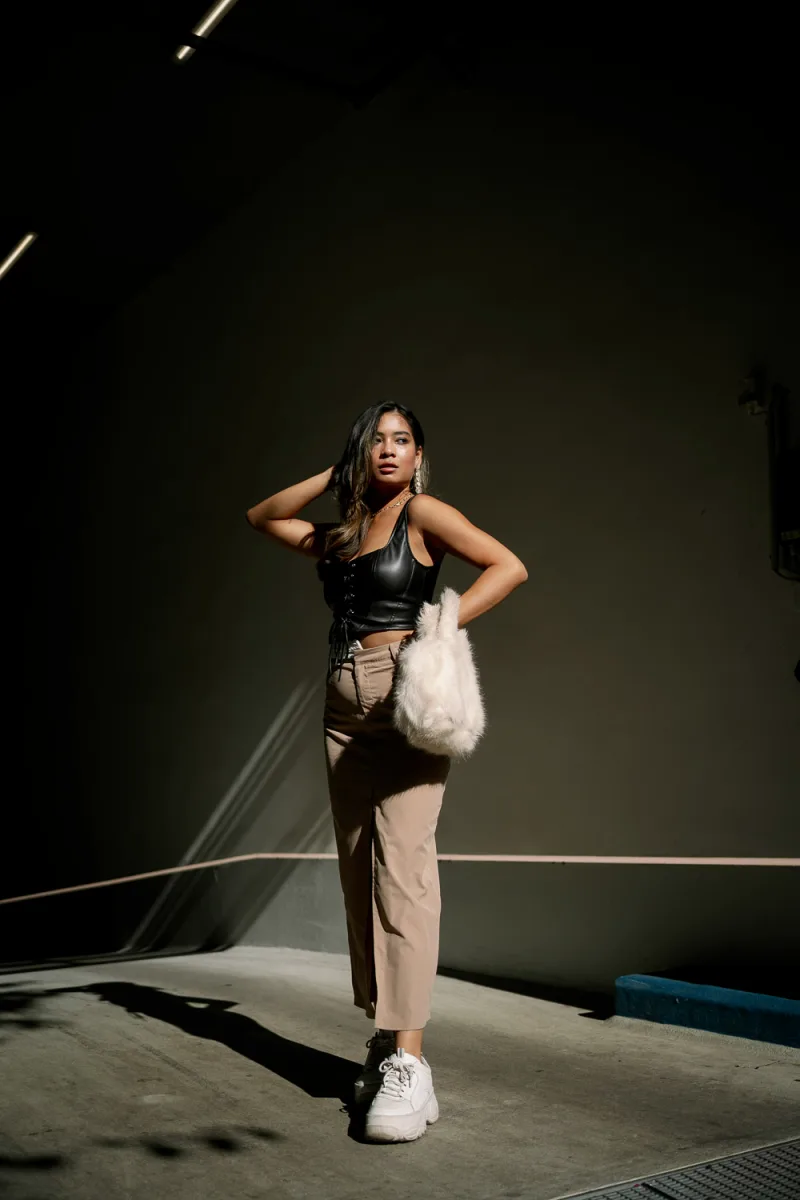 Woman in black leather top and beige pants with white fur bag on dark background