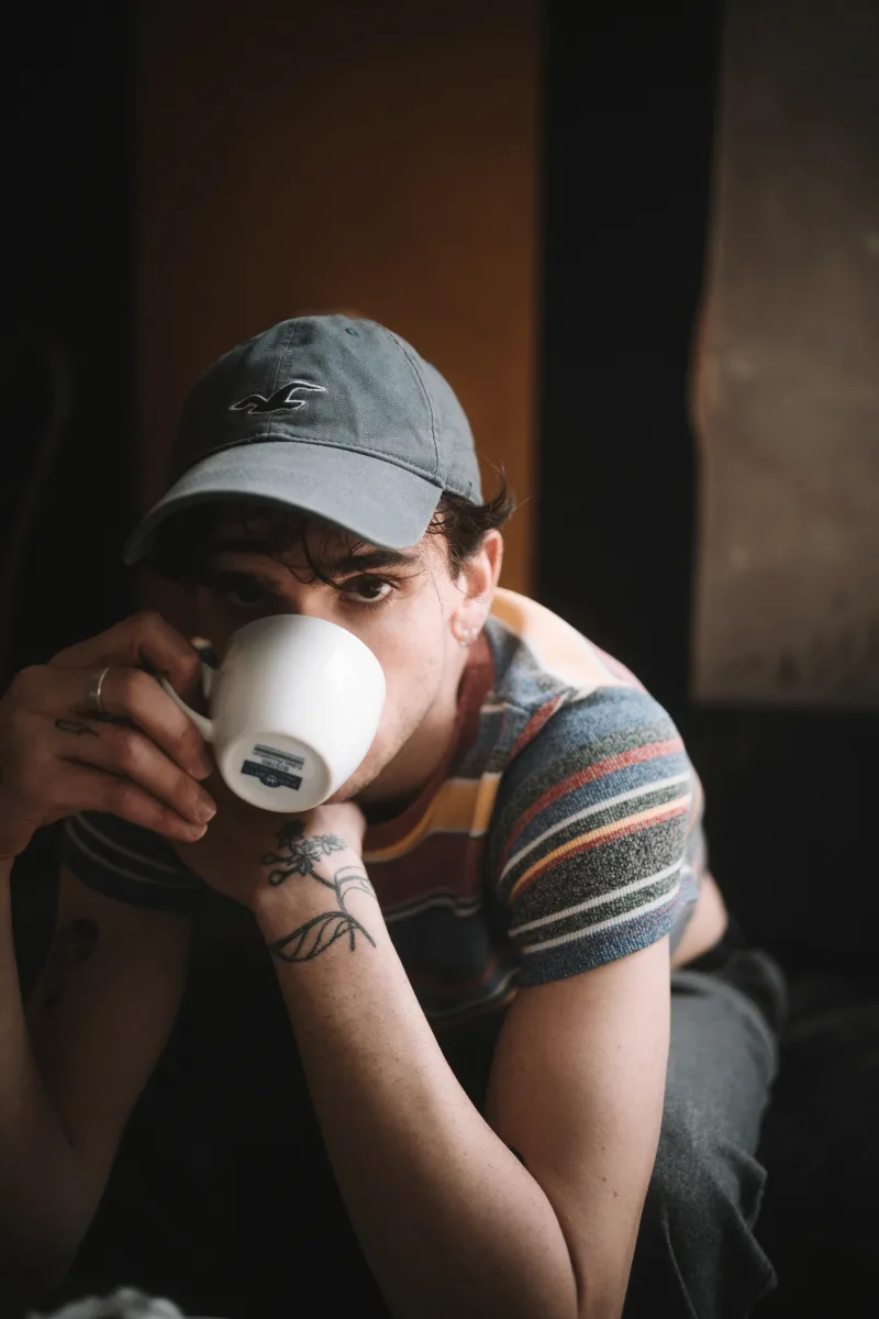 Man in blue cap holding white mug with both tattooed hands drinking