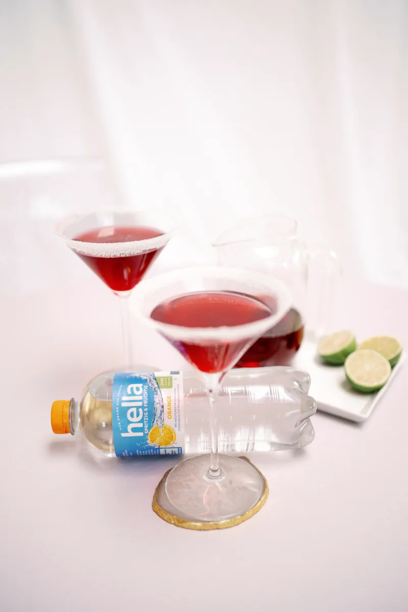 Red cocktails with salt rims, lime slices, and sparkling water bottle on white surface