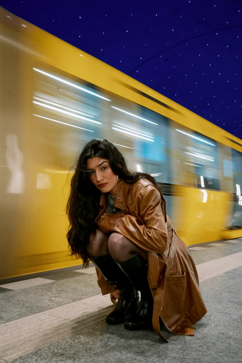 Woman in tan leather jacket crouching by passing yellow train at night, urban