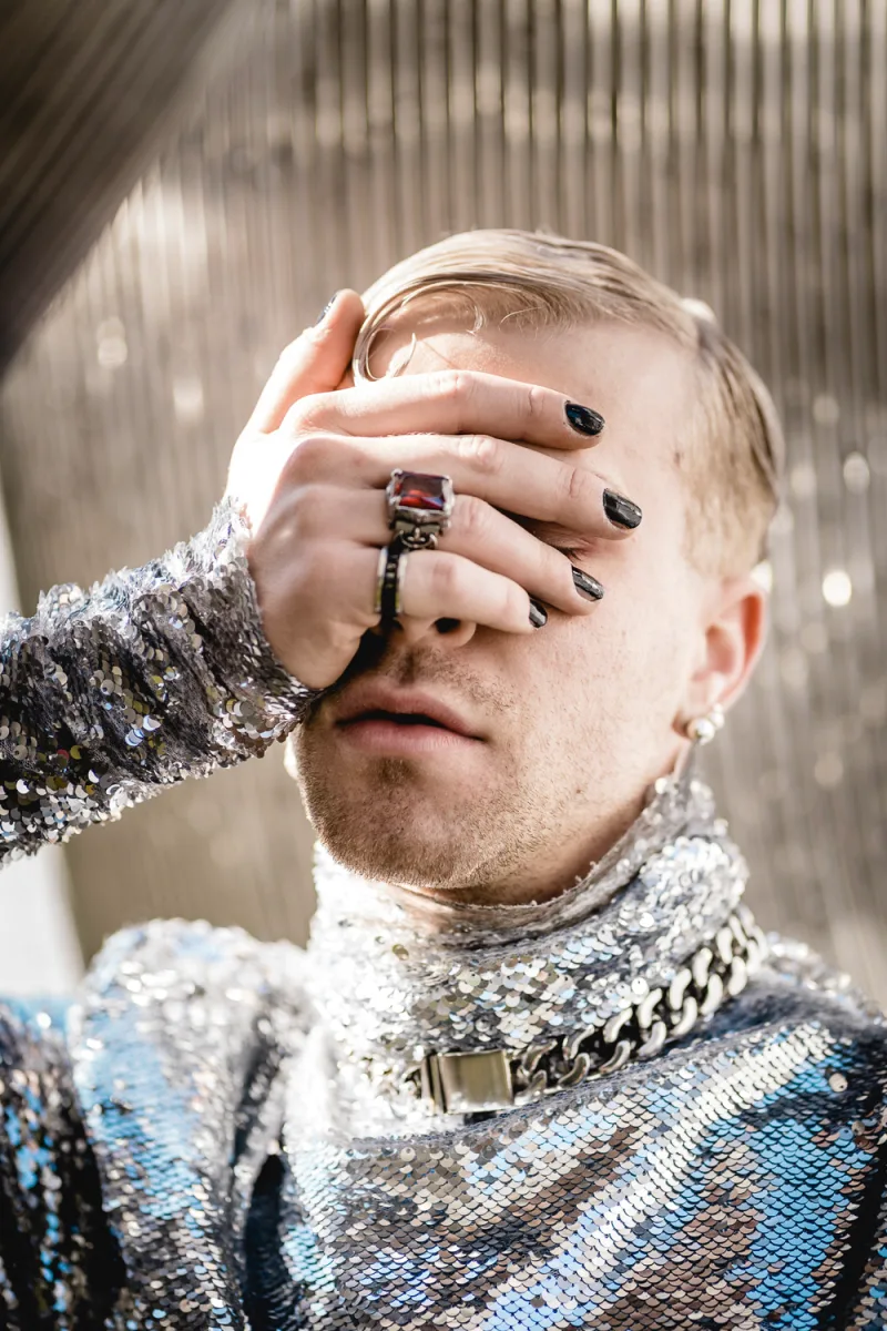 Person with blonde hair in silver sequined outfit holding face with hands