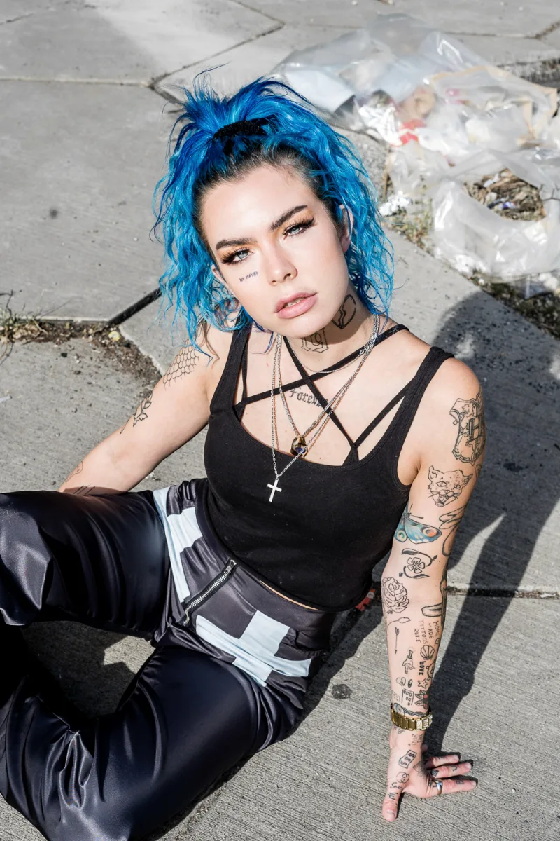 Woman with bright blue curly hair in black top with arm sleeve tattoos