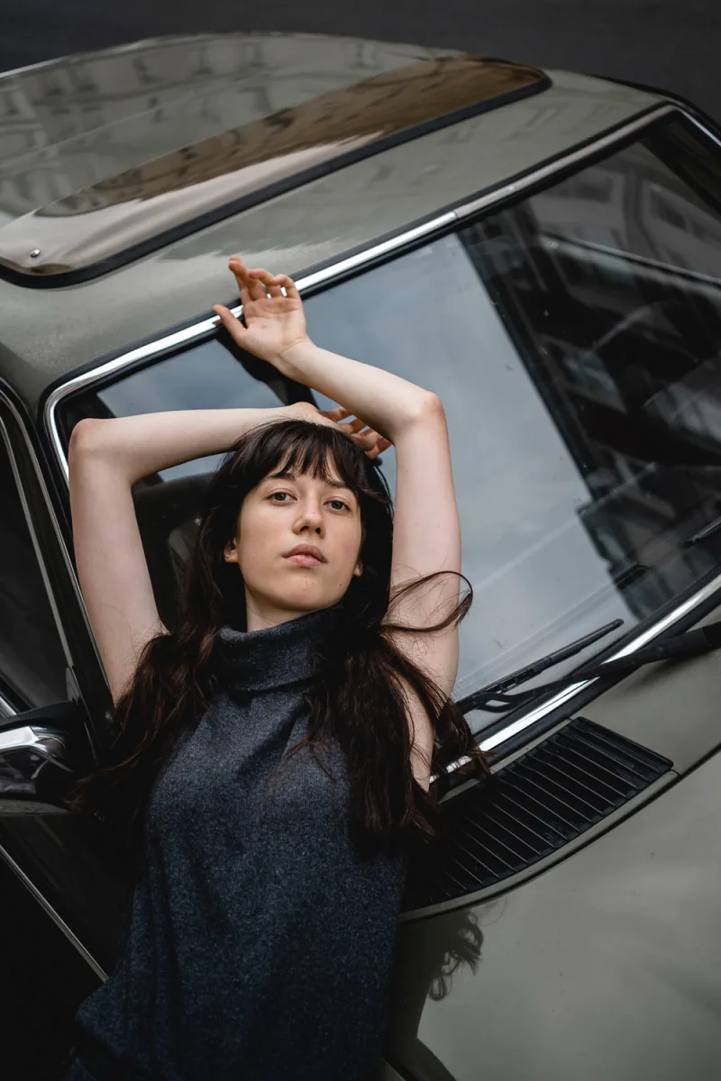 Woman with dark hair and bangs with arms raised looking up from car window