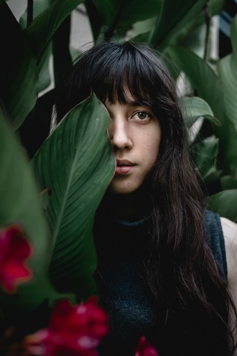 Woman with dark hair partially hidden among green plant leaves with red flowers