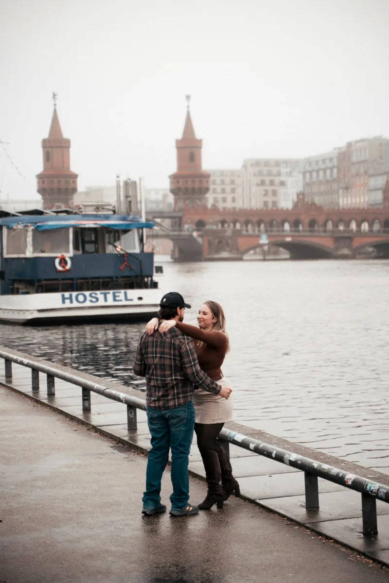 Couple standing by waterfront railing with historic brick bridge towers and boat behind