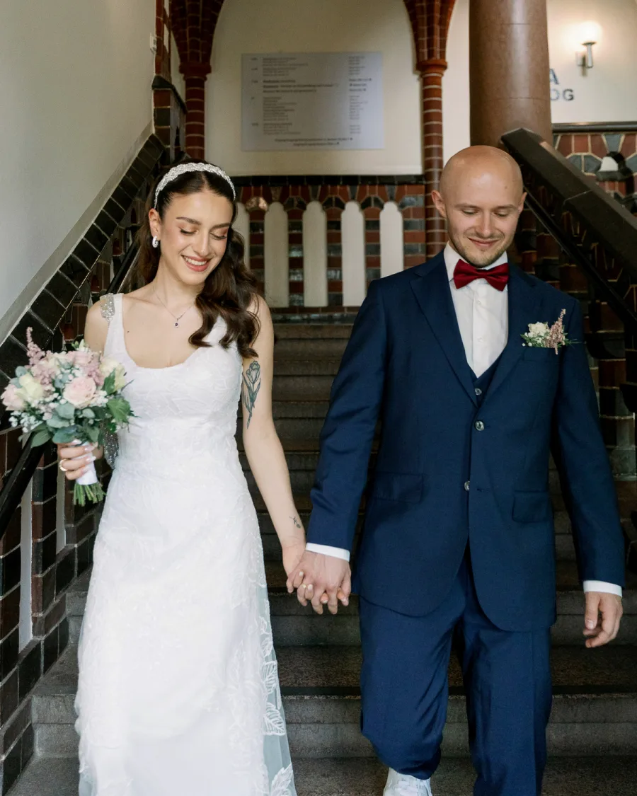 Bride in white lace dress and groom in navy suit on interior staircase