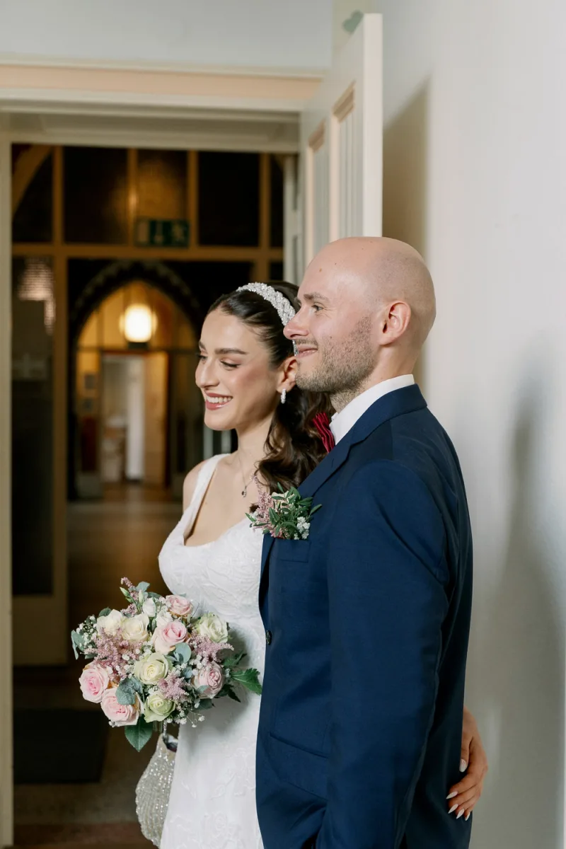Bride in white lace gown with groom in navy suit holding pale pink flower bouquet