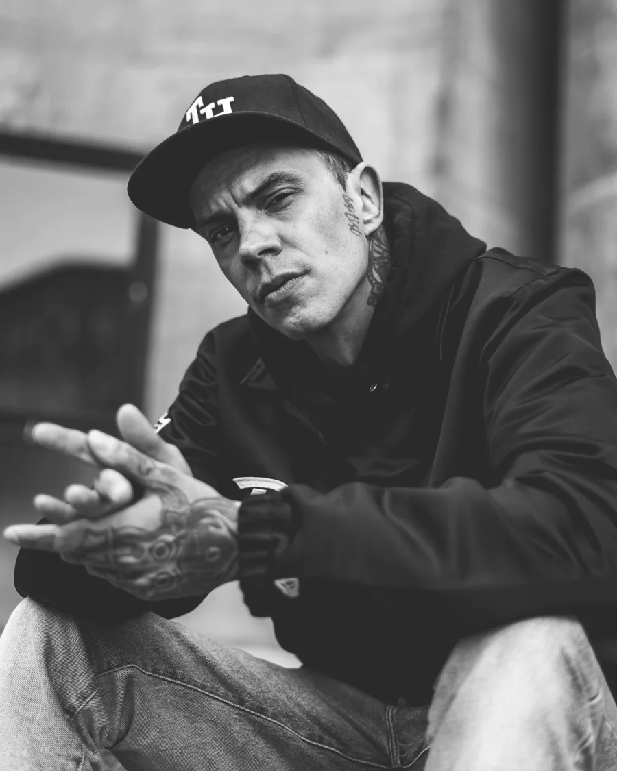 Man in black cap and jacket with tattooed hands looking downward black and white