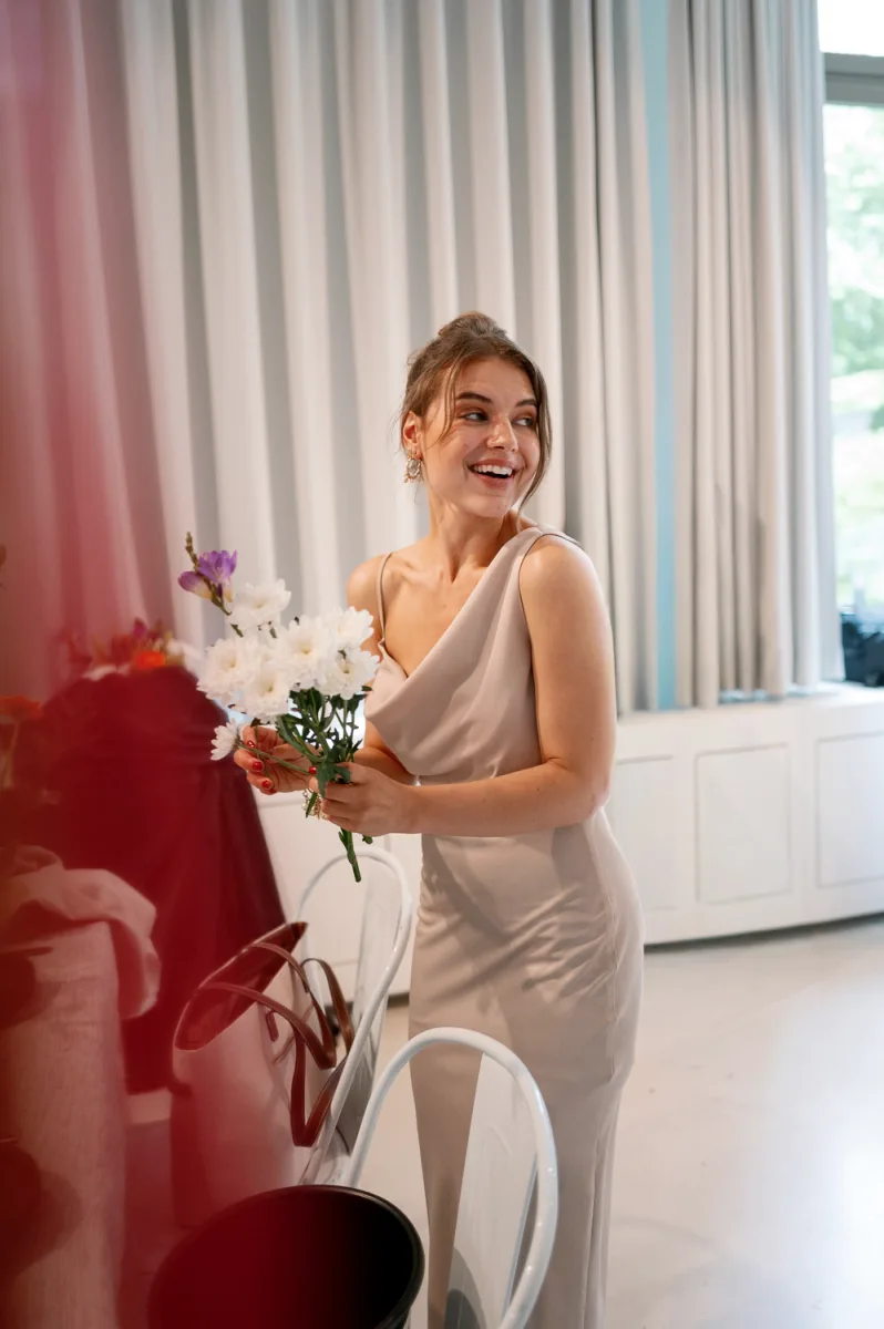 Woman in beige dress smiling while holding white and red flower bouquet indoors