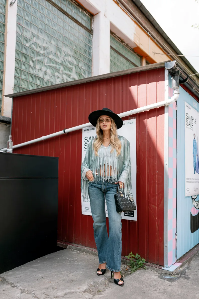 Woman with blonde wavy hair in black cowboy hat, fringe vest near red building