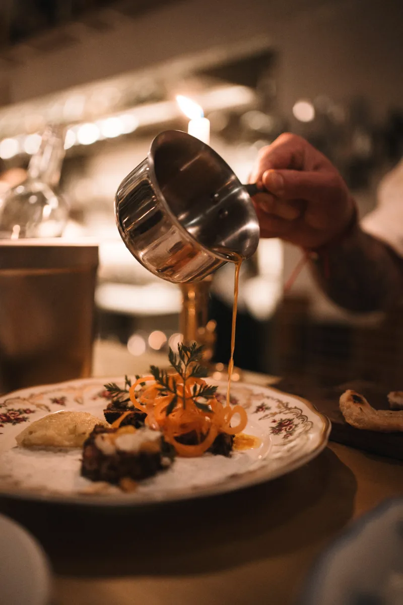 Hand pouring golden sauce over artfully plated dish with garnish and lighting