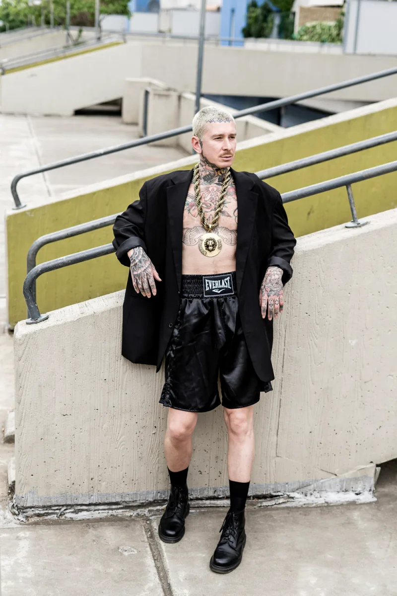 Heavily tattooed man with platinum hair posing against industrial metal staircase