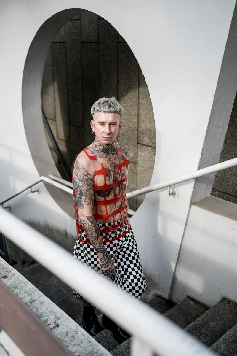 Person with platinum blonde hair and tattoos in red and black checked pants