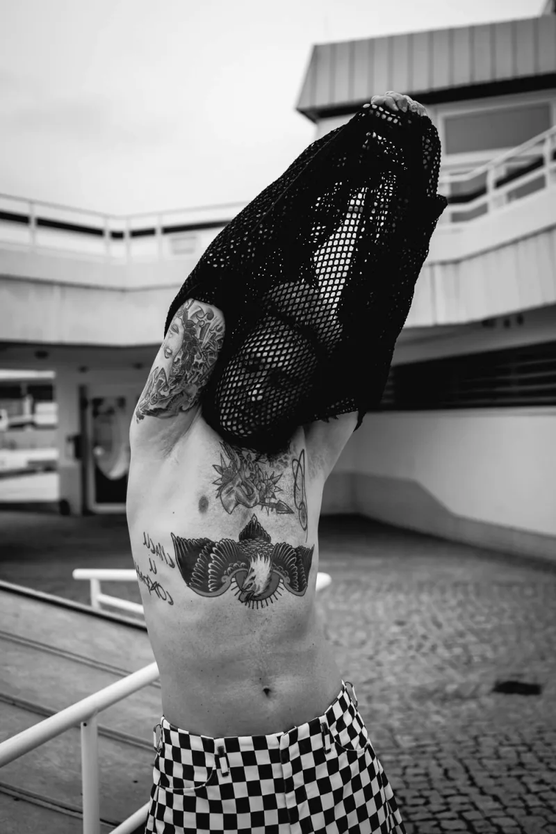 Shirtless tattooed man with mesh overlay covering face against parking structure