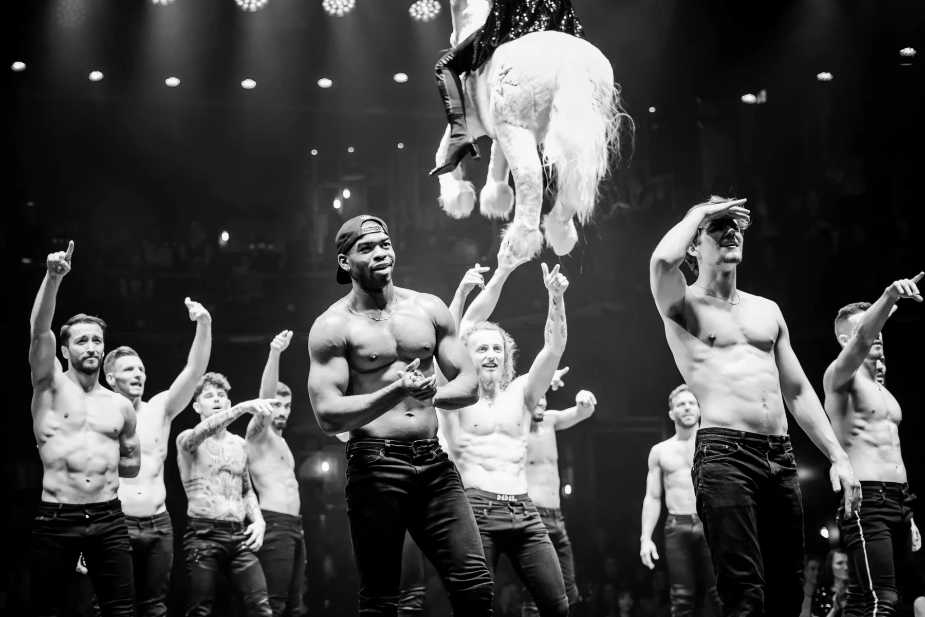 Group of shirtless male performers on stage under bright theatrical lighting