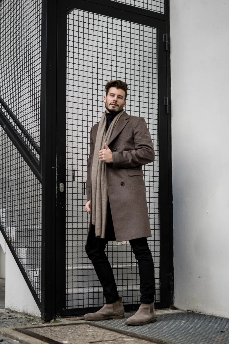 Man in brown camel coat and black jeans posing at black metal mesh gate