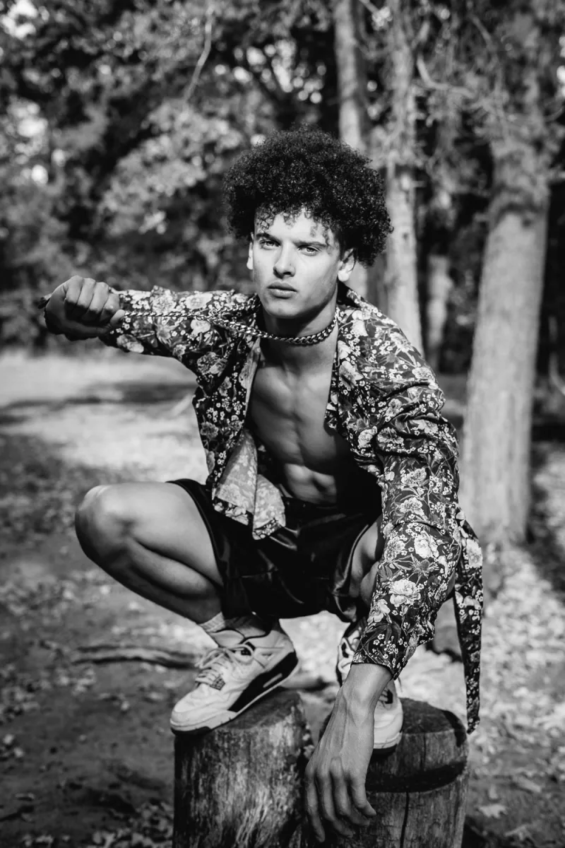 Man in colorful patterned shirt crouching on wooden log in dense green forest