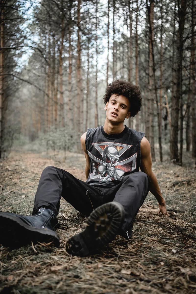 Young person in black graphic shirt sitting pensively in forest surrounded by tall trees
