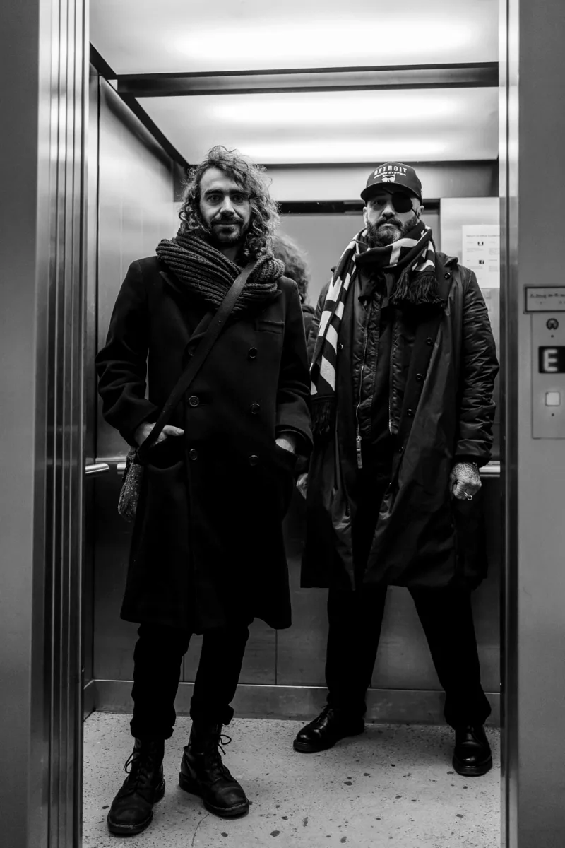 Two men in black and white one with curly hair one with hat