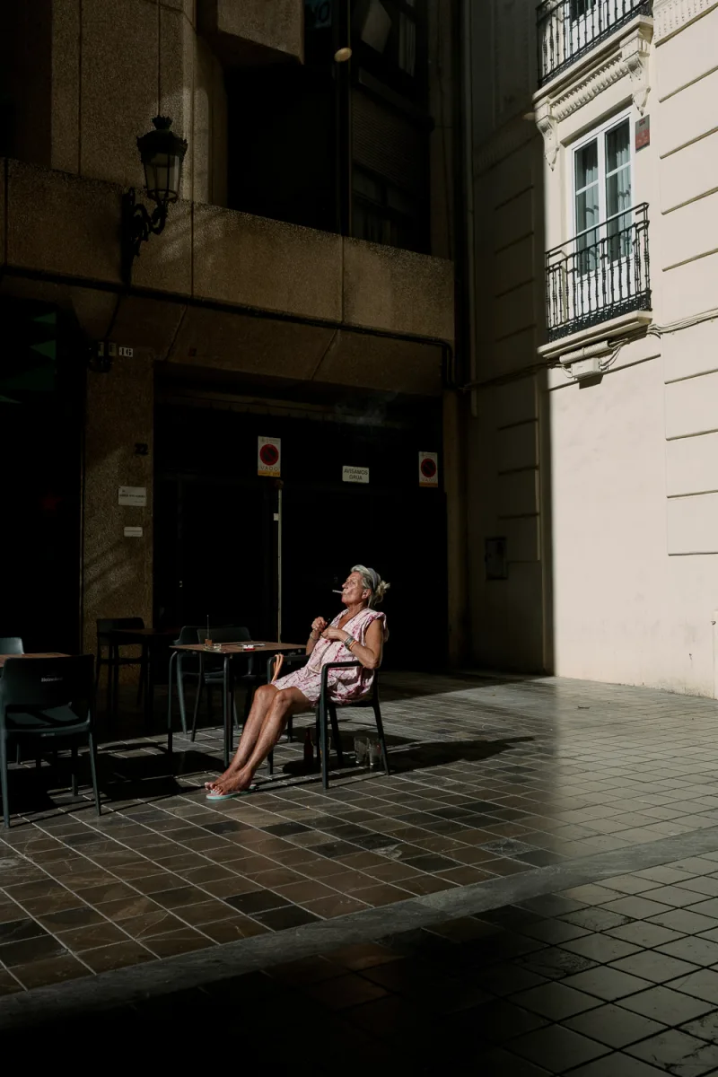 Woman seated at outdoor table under historic architecture, smoking, warm lighting