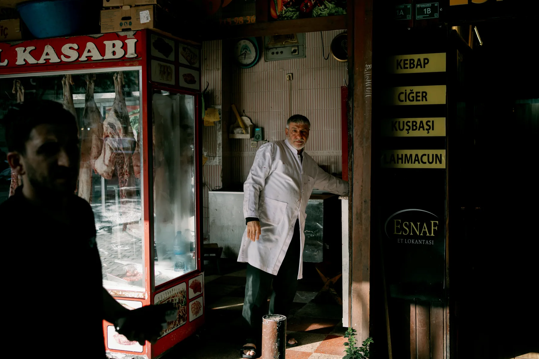Man in white coat standing in traditional meat shop with hanging carcasses