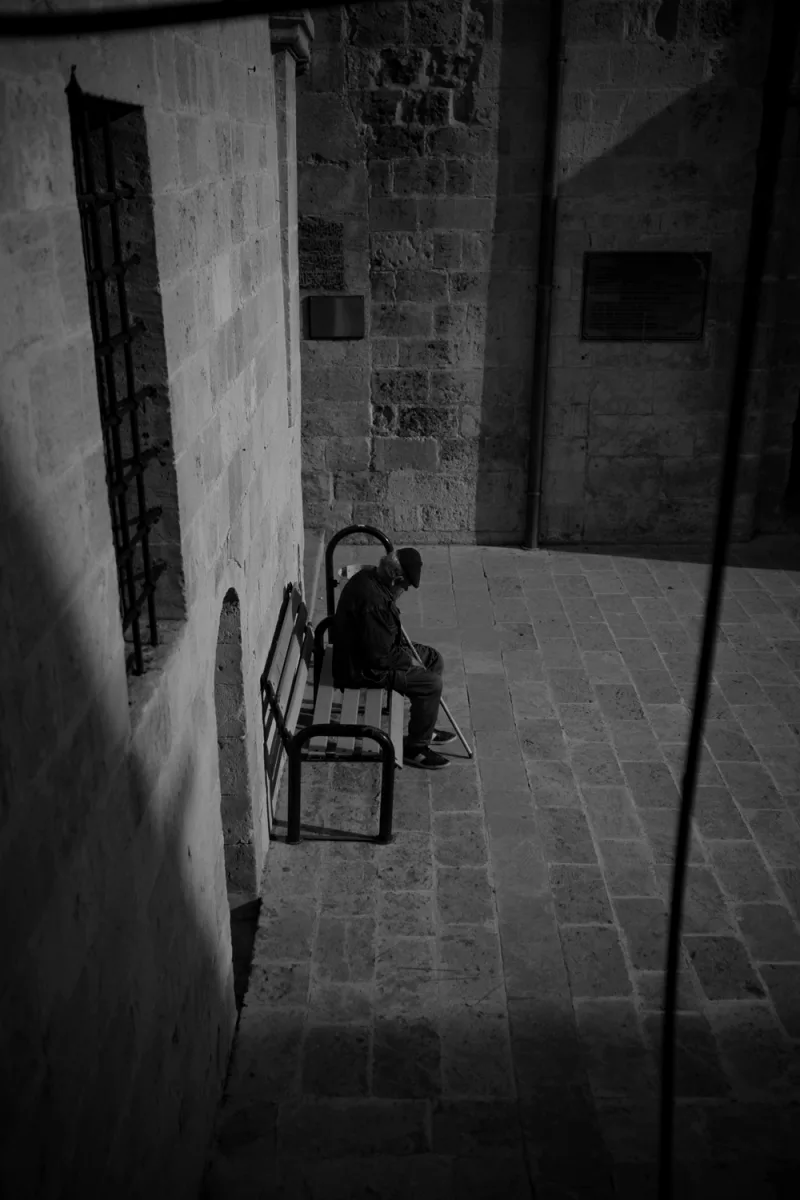 Person in dark coat sitting alone on wooden bench in historic stone courtyard with arches