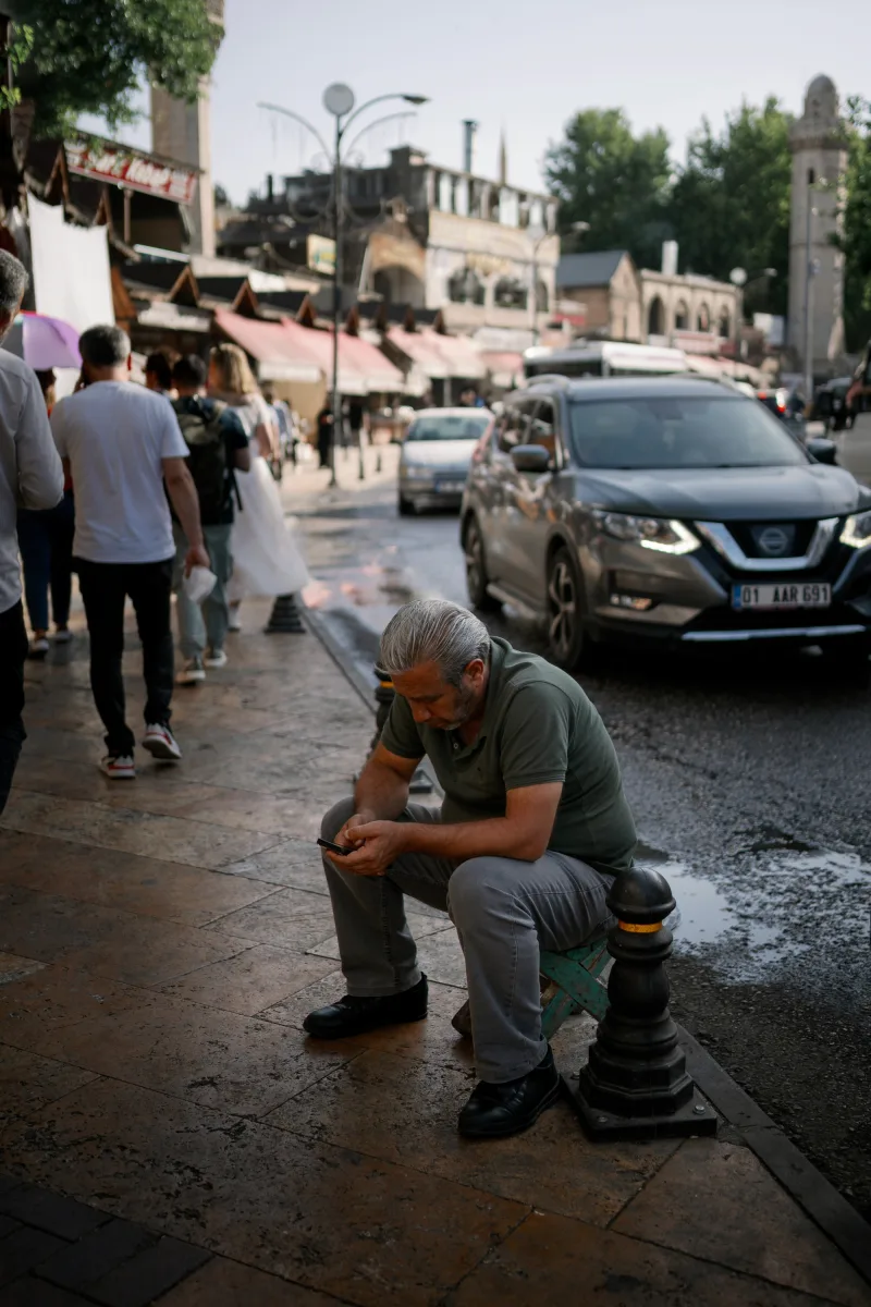 Older man with grey beard sitting on street step amid bustling marketplace