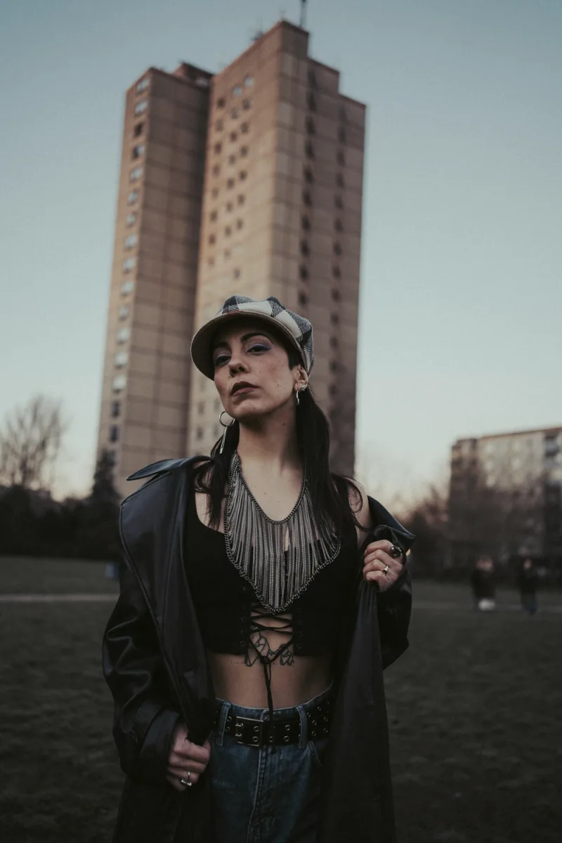 Woman in dark urban clothing with cap and earrings against tall brick buildings