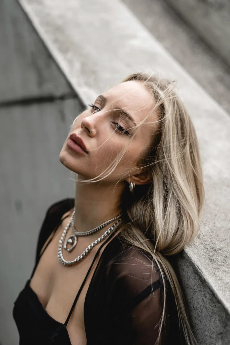 Woman with blonde hair tilted back against concrete wall, pearl chain necklace visible