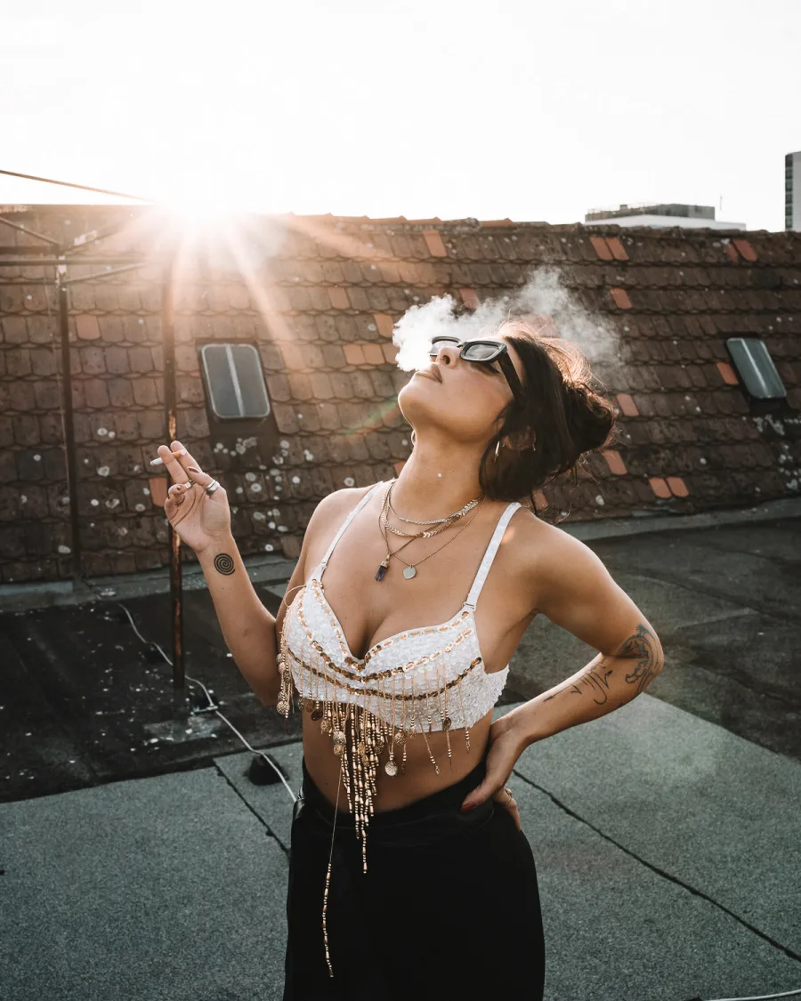 Woman in beaded white crop top on rooftop at golden hour, exhaling smoke