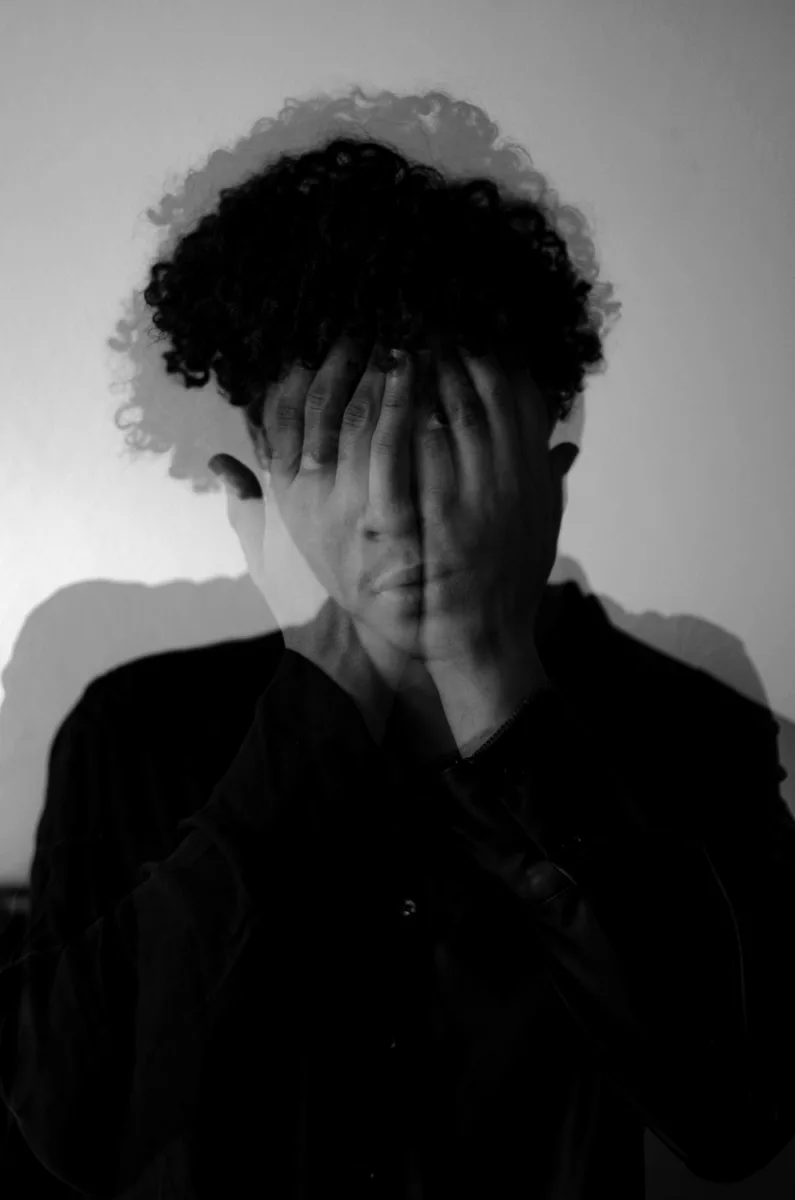 Double-exposure black and white portrait, man with curly hair covering face with hands
